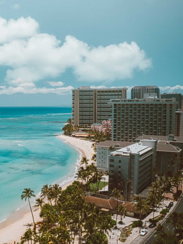 Coastal view featuring buildings along a sandy beach and turquoise water under a blue sky.